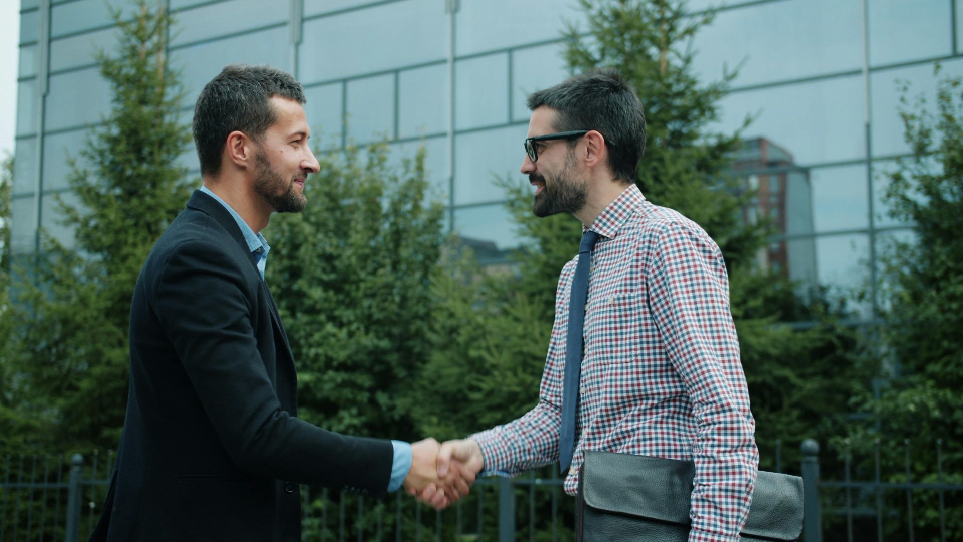 Zwei Männer schütteln sich vor einem modernen Bürogebäude die Hände – ein Symbol für Geschäftspartnerschaft.