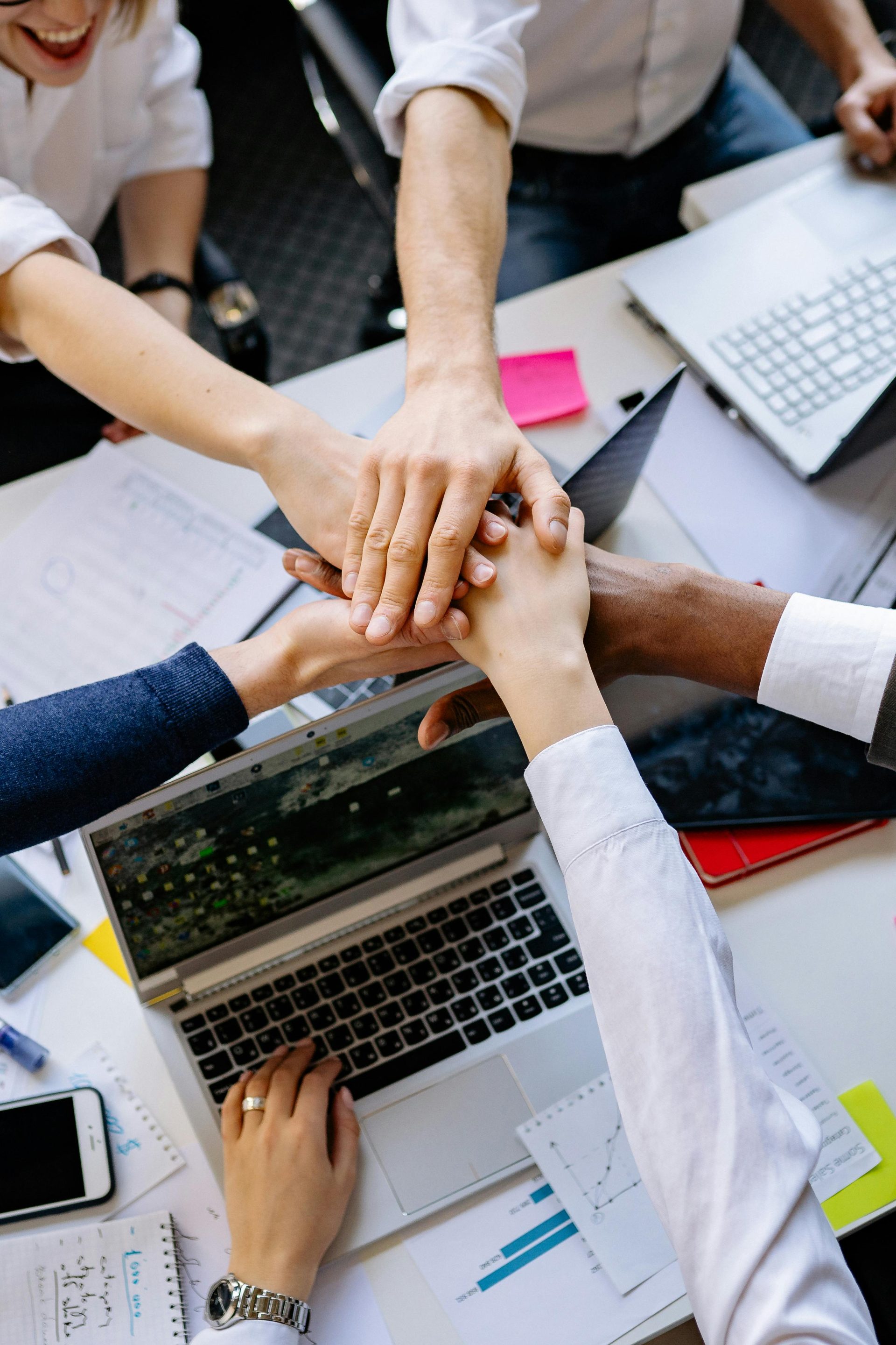 Eine Gruppe von Fachleuten aus verschiedenen Bereichen legt die Hände übereinander auf einen Tisch, um Teamwork und Zusammenarbeit zu symbolisieren.